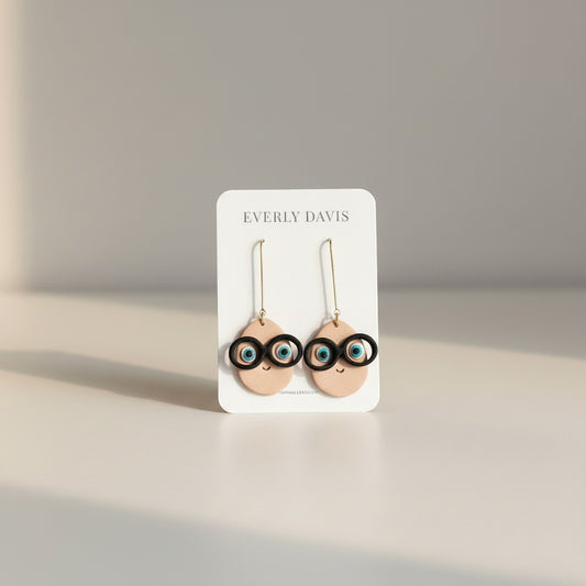 Everly Davis tan clay egg-shaped earrings with black glasses and blue eyes, dangling from gold hooks.