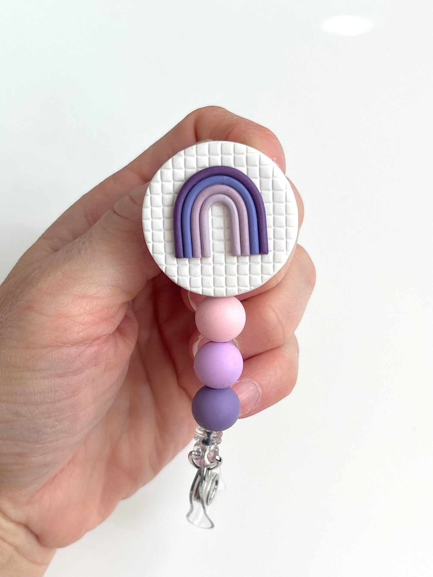 Everly Davis handmade badge reel with a white textured circle, purple rainbow charm, and pink, lavender, and purple silicone beads.