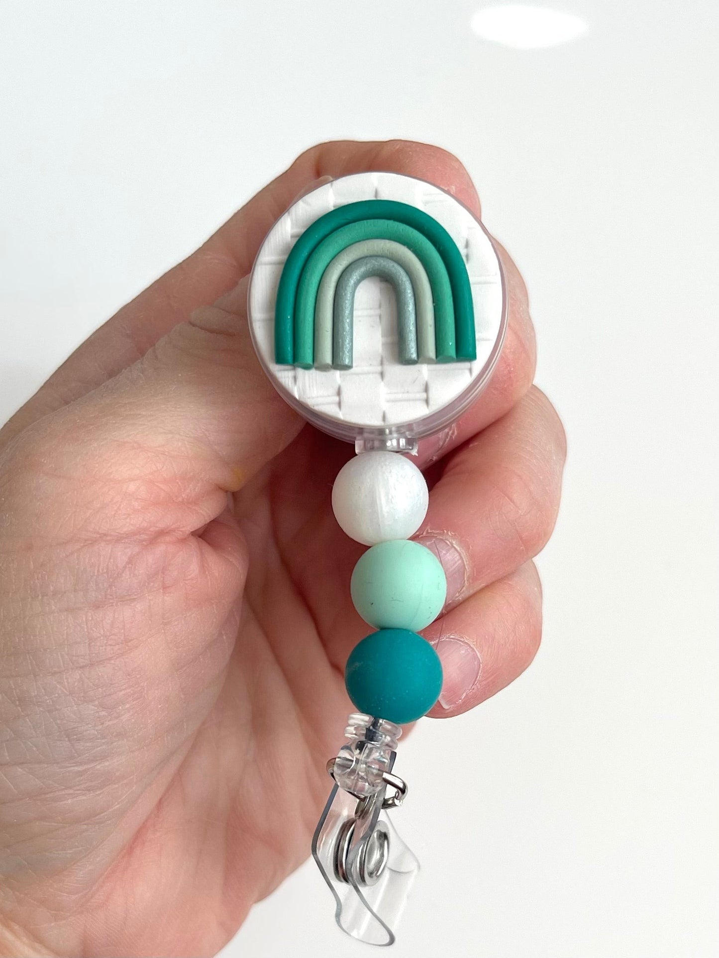 Everly Davis handmade beaded badge reel with a white textured background and a green rainbow design. Features white, mint, and teal silicone beads.