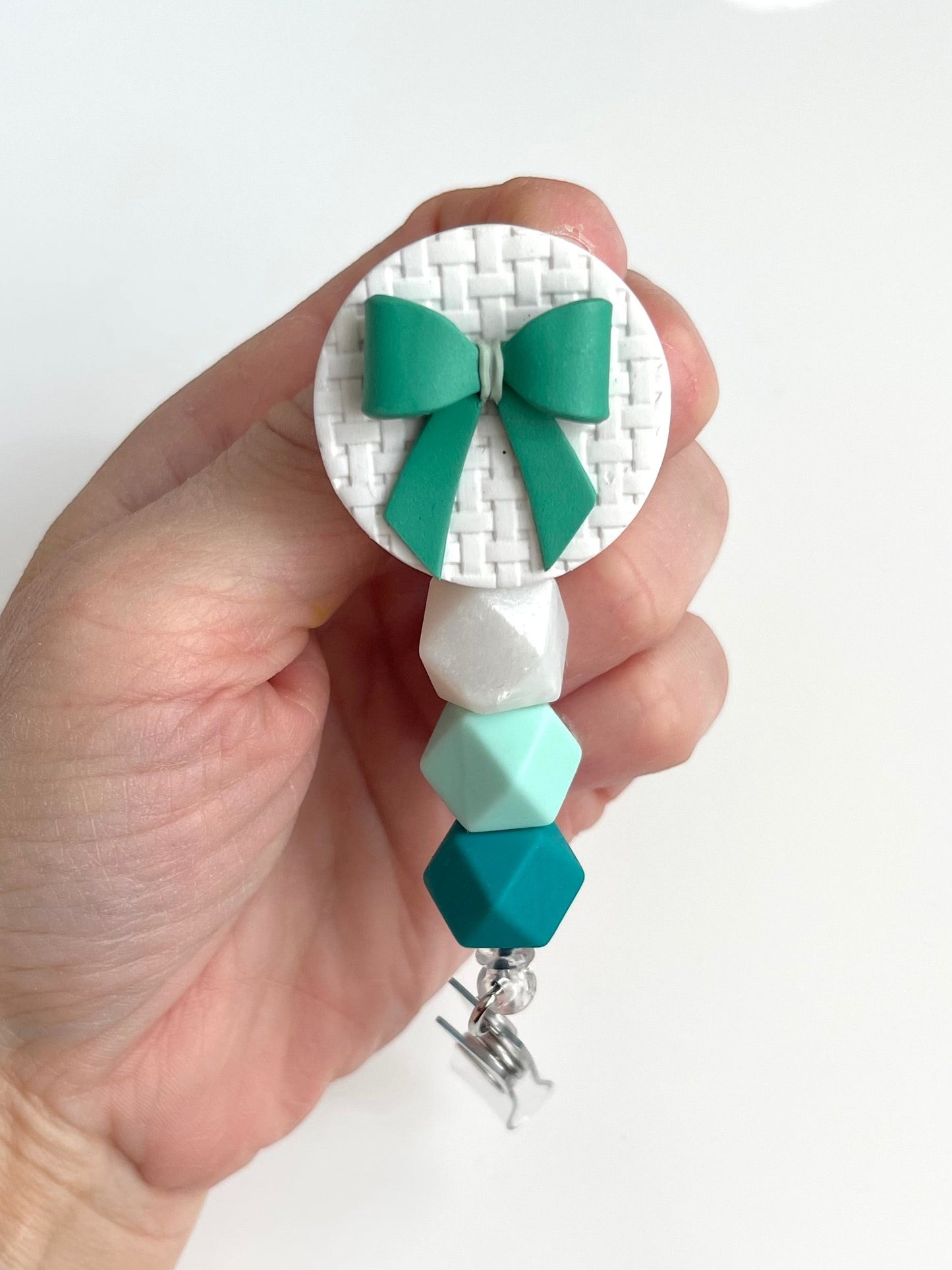 Everly Davis handmade teal bow badge reel with white textured circle and teal, mint, and white silicone beads.